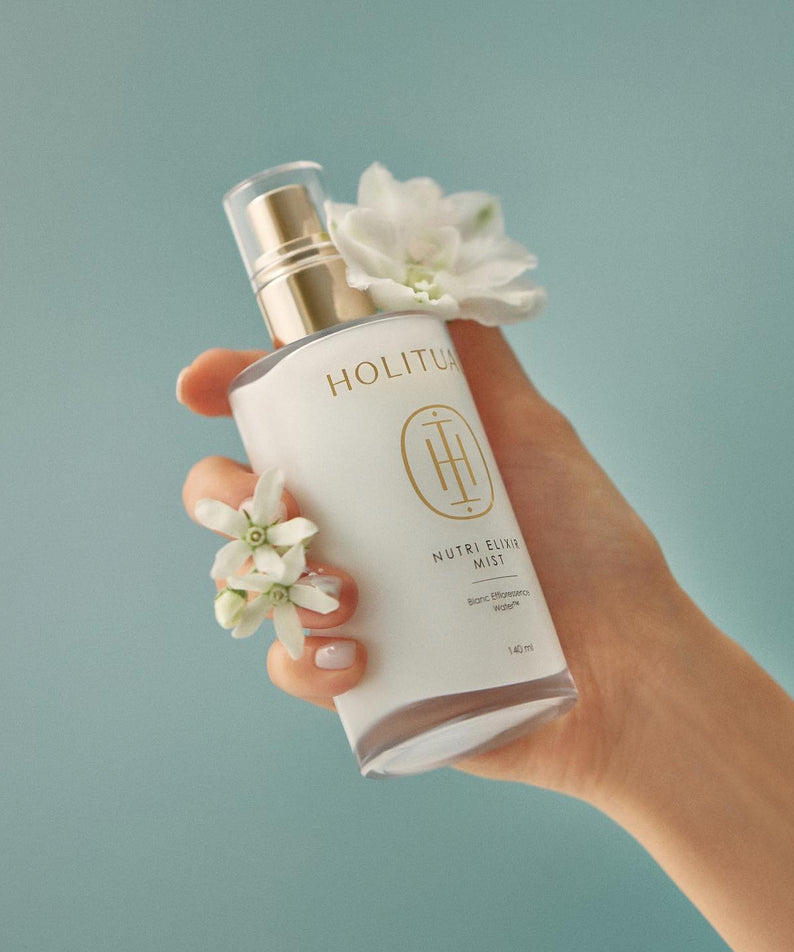 Main tenant HOLITUAL  Elixir Mist 140ml, flacon blanc et fleurs sur fond bleu, marque HOLITUAL en évidence