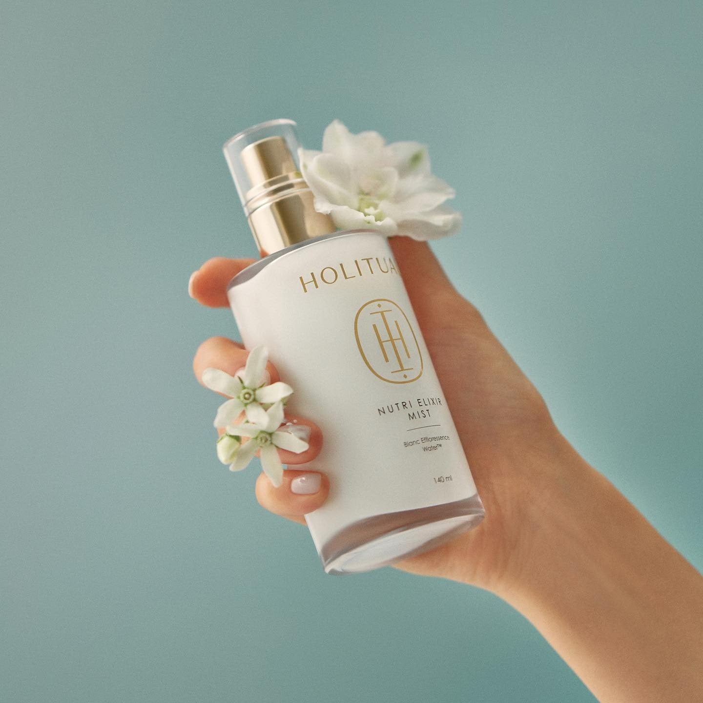 Main tenant HOLITUAL  Elixir Mist 140ml, flacon blanc et fleurs sur fond bleu, marque HOLITUAL en évidence