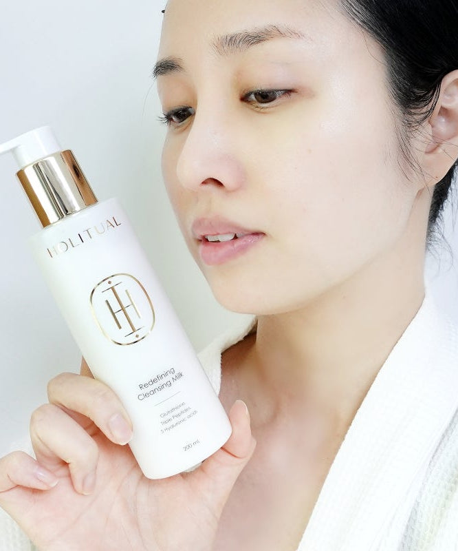 HOLITUAL Redefining acleansing Milk 200ml, femme tenant le flacon blanc et or, routine de soin visage sur fond clair