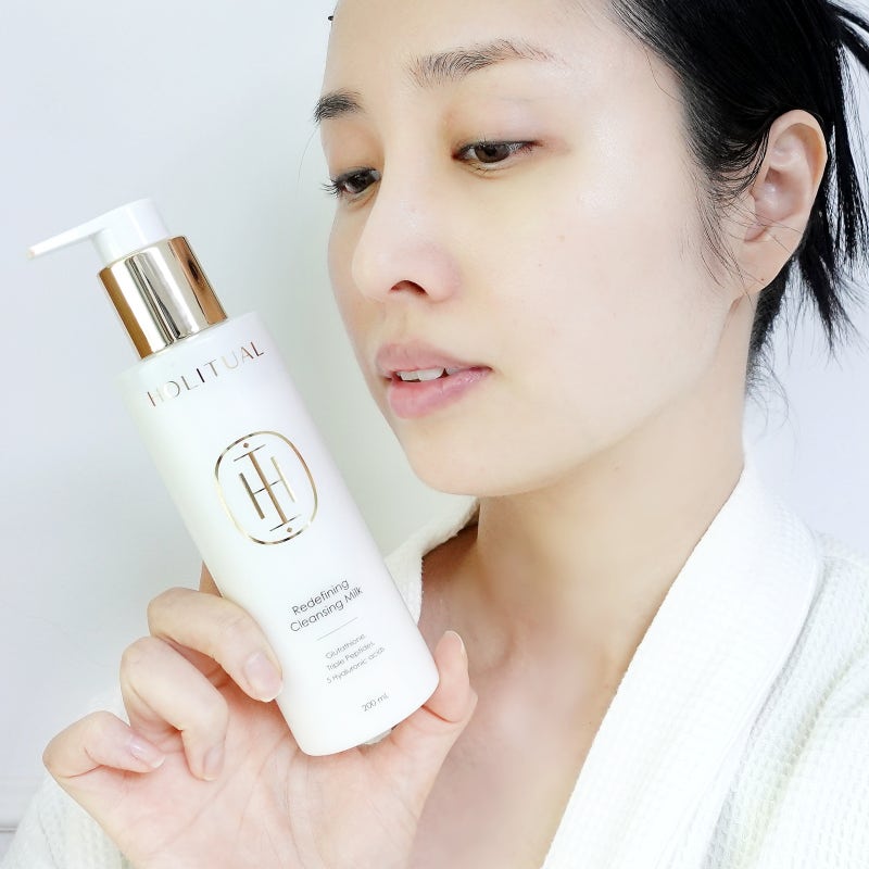 HOLITUAL Redefining acleansing Milk 200ml, femme tenant le flacon blanc et or, routine de soin visage sur fond clair