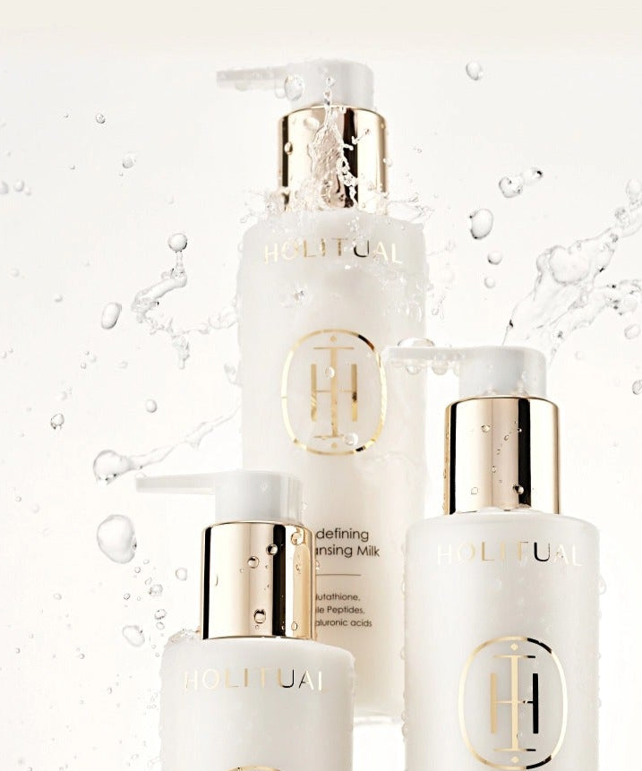Ensemble de soins de la peau HOLITUAL Redefining acleansing Milk 200ml avec pompe dorée, entouré de gouttes d'eau élégantes.