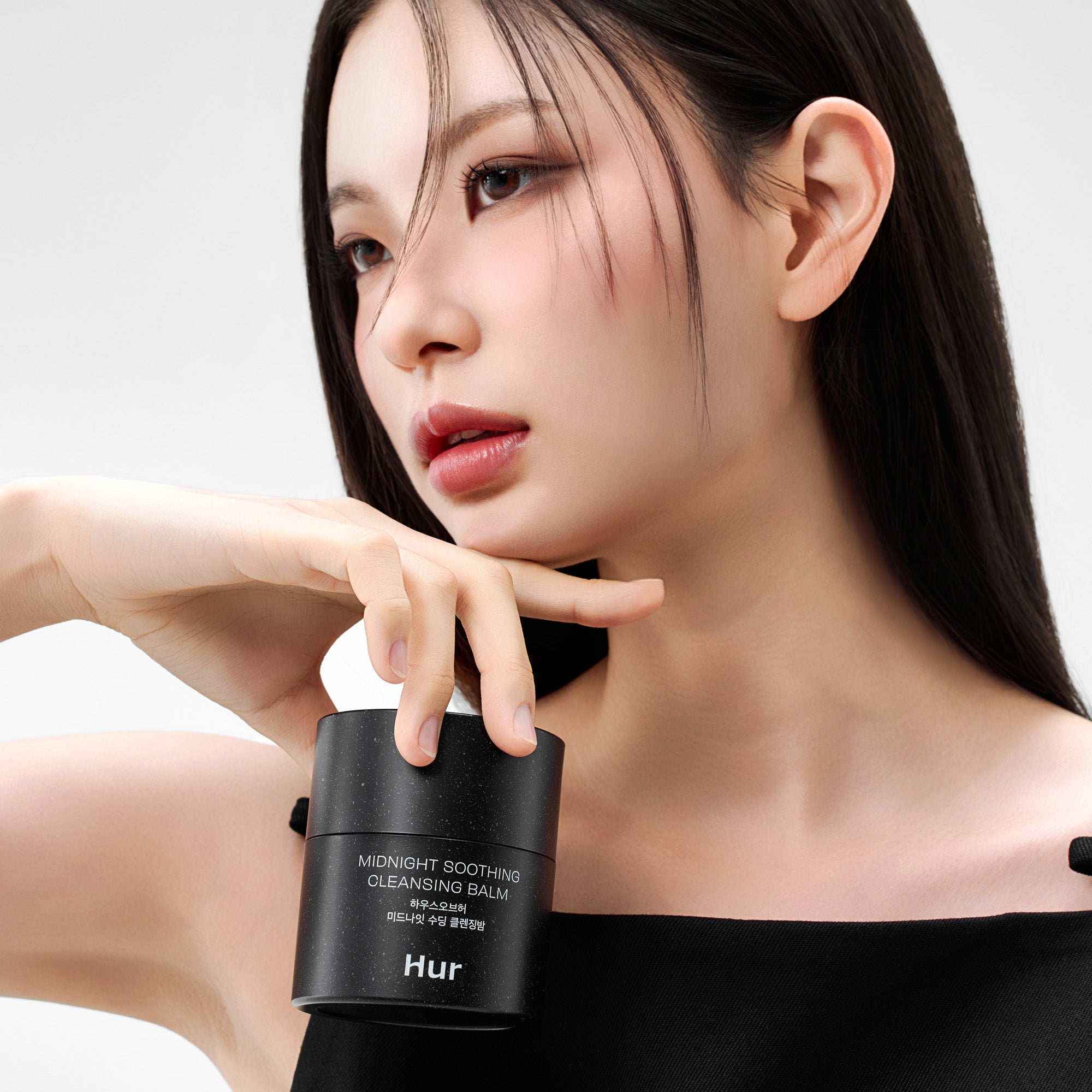 Décris l’image en français : HOUSE OF HUR Midnight Soothing Cleansing Balm HUR, femme en soin, pot noir sur fond clair.