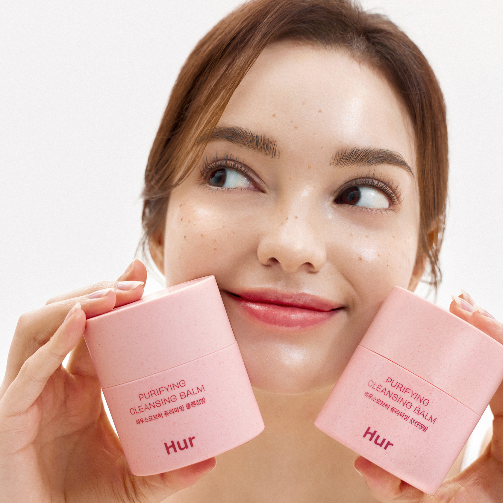Décris l'image en français : HOUSE OF HUR Purifying Cleansing Balm, femme tenant deux baumes roses contre fond blanc.