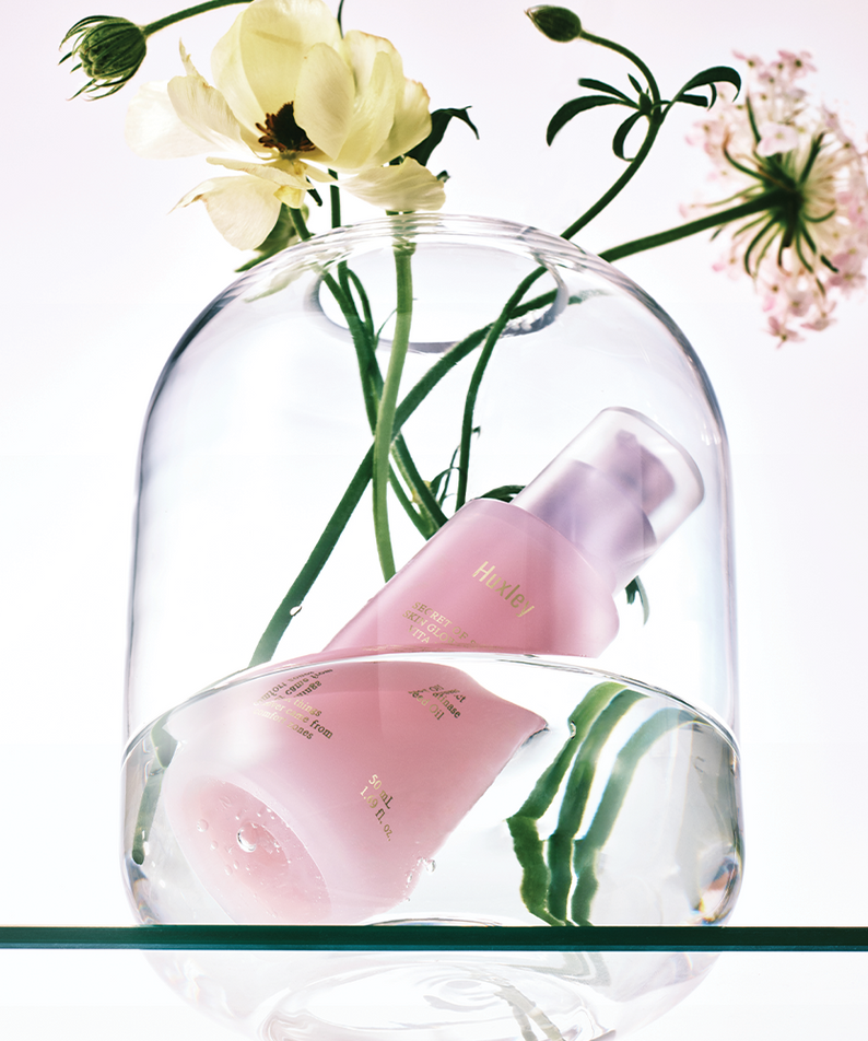HUXLEY Skin Glow Essence Vita Radiance en bouteille rose avec fleurs en arrière-plan sous un verre transparent.