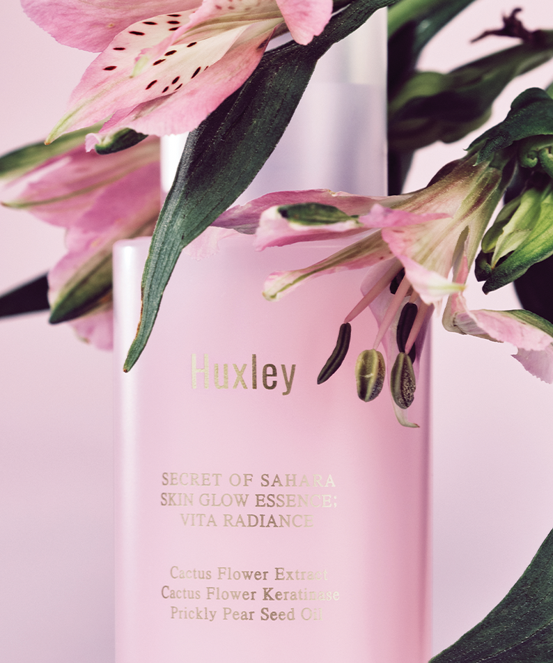 HUXLEY Skin Glow Essence Vita Radiance, soin éclat avec extrait de fleur de cactus et huile de figue de barbarie, flacon sur fond floral.