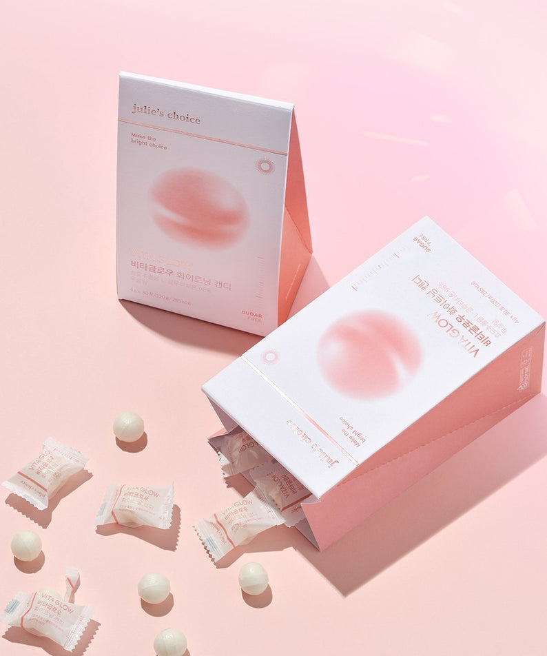 Boîtes de JULIE'S CHOICE Vita Glow Whitening Candy sur fond rose avec bonbons éparpillés. Produit éclaircissant pour la peau.