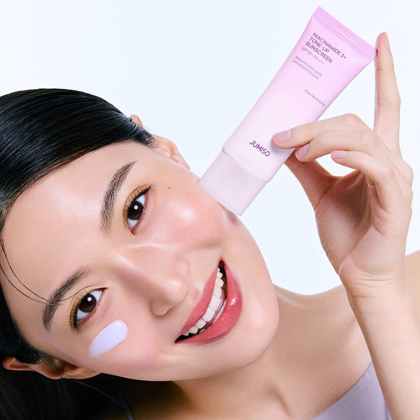 Décris l'image en français : JUMISO  Niacinamide 3 Plus Tone-Up Sunscreen 40ml, tube rose sur peau, main et visage.