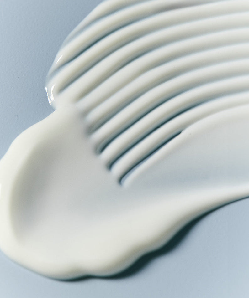 Texture onctueuse du JUMISO Hyaluronic Acid Water Plumping Moisture Sun Serum 40ml, capture de la crème étalée, fond bleuâtre.