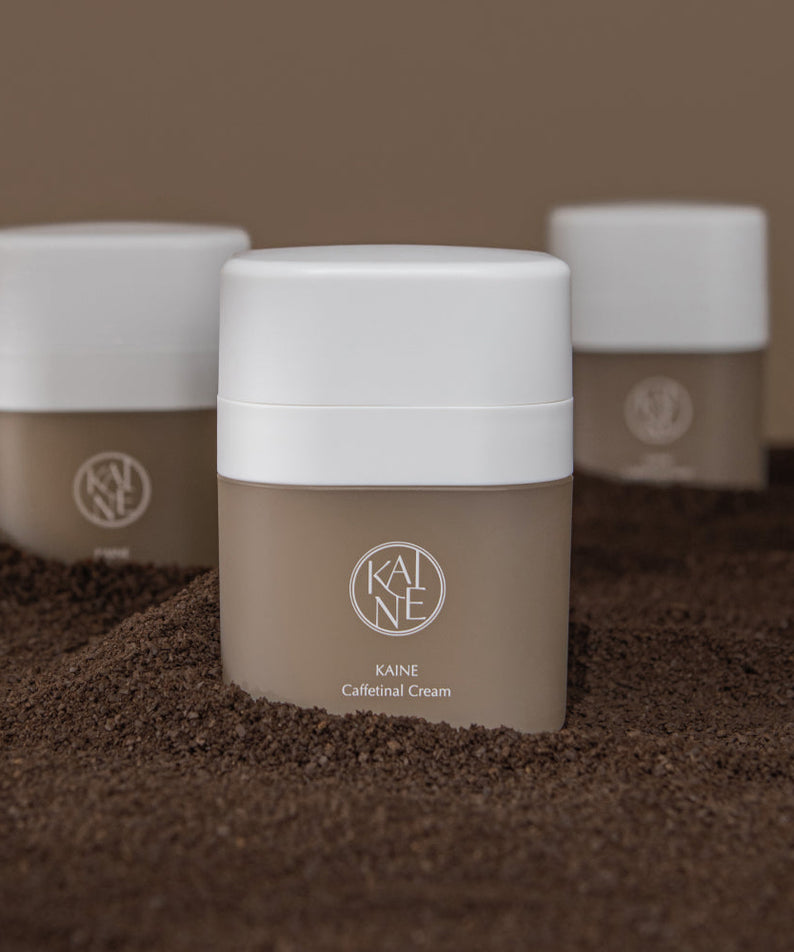 Trois pots de KAINE Caffetinal Cream 50ml posés sur un lit de café moulu, mettant en avant le design moderne du produit.