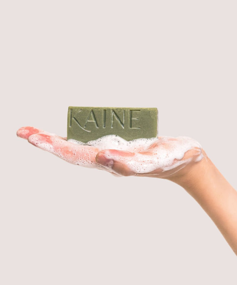 Savon KAINE Rosemary Acne Mildly Acidic Cleansing Bar 100g mousseux tenu par une main, nettoyant doux pour peaux à tendance acnéique.