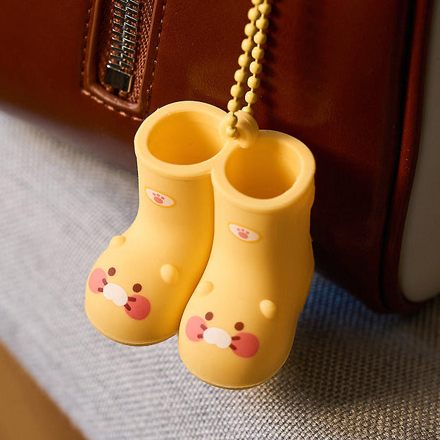 KAKAO FRIENDS Rain boots keychain, porte-clés bottes de pluie KAKAO FRIENDS jaune, sur fond tissu, près d’un sac marron.