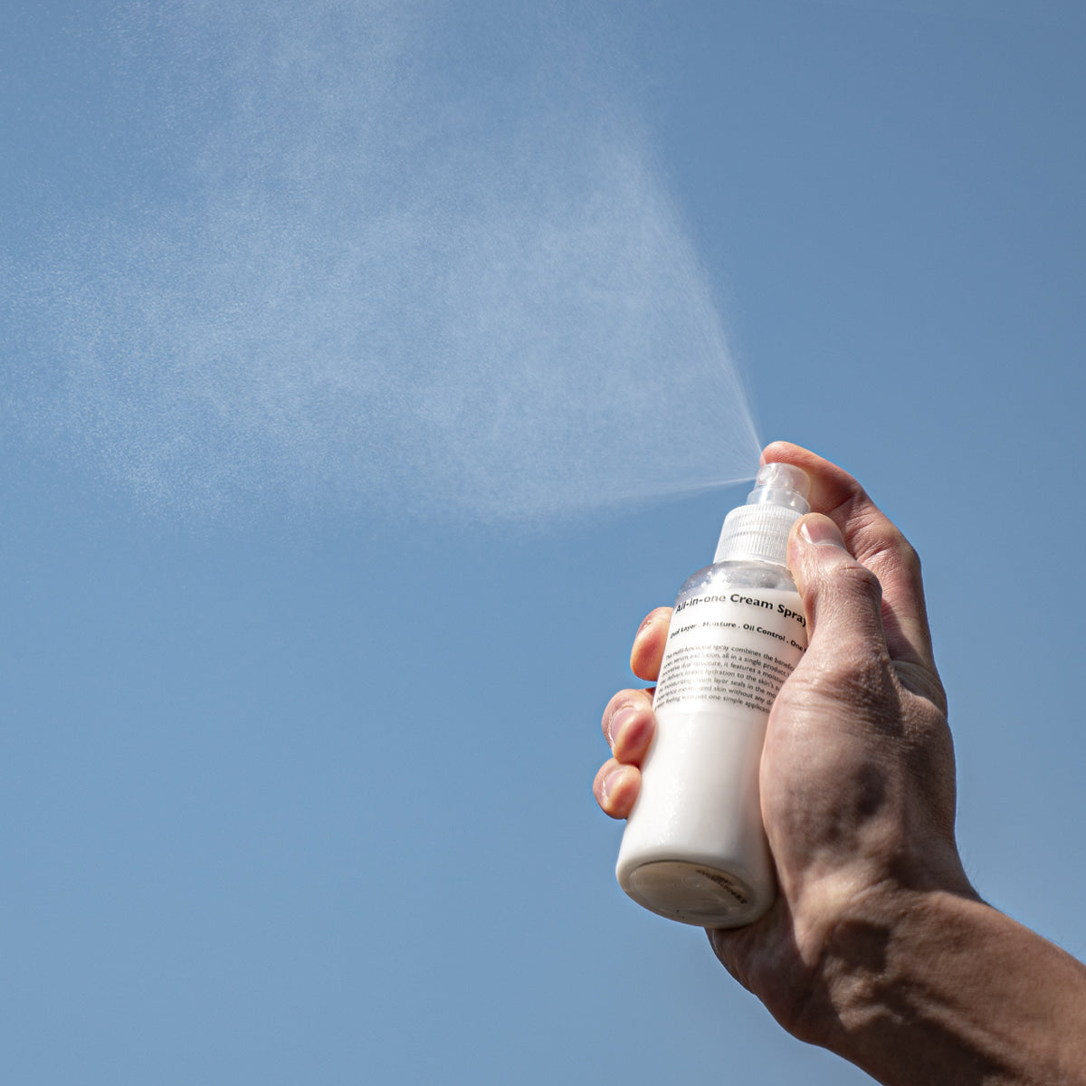 LAZY SOCIETY All-in-One Cream Spray 120ml, main tient le spray et projette une brume de crème, fond bleu.