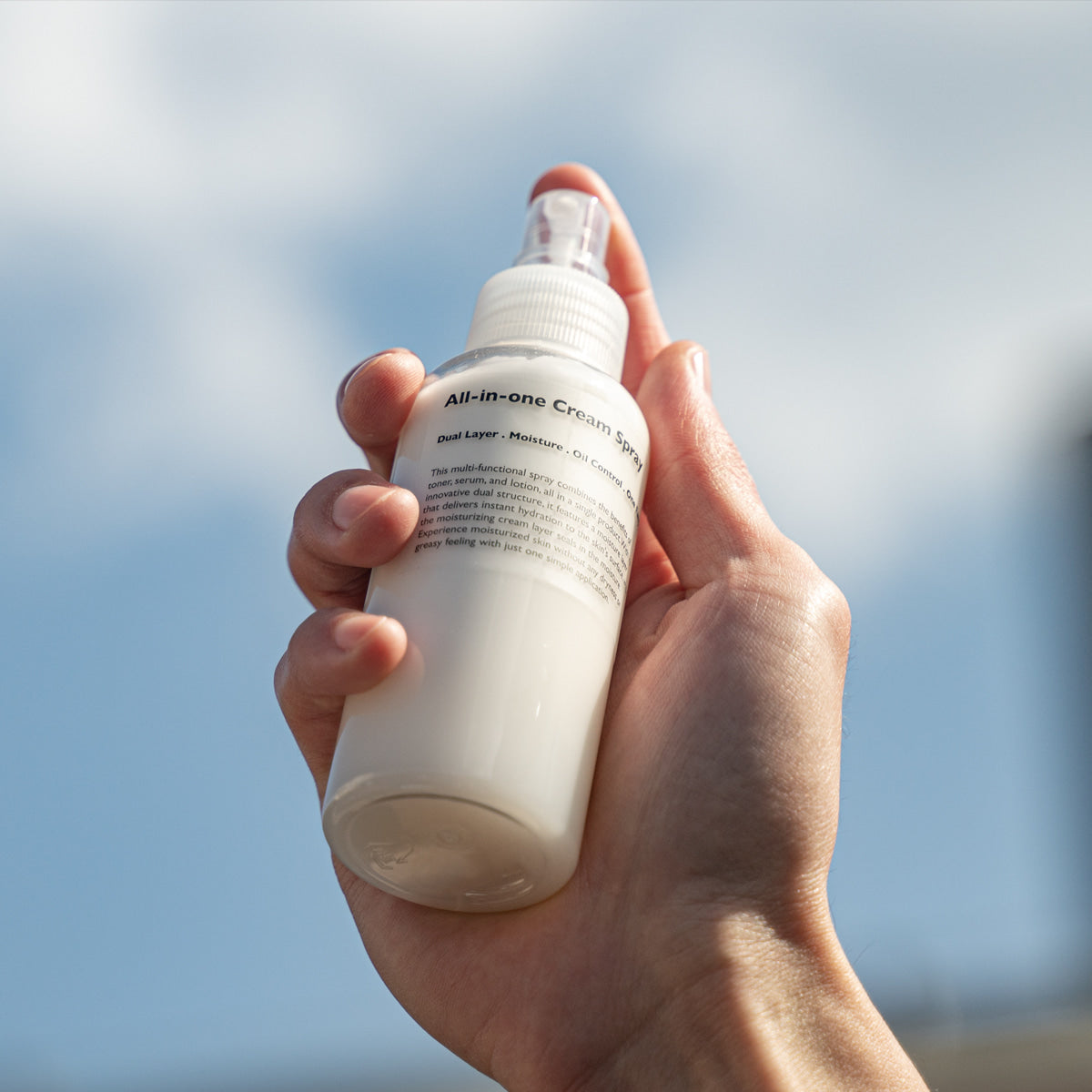 LAZY SOCIETY All-in-One Cream Spray 120ml : flacon crème all-in-one tenu à la main, fond ciel bleu, vue rapprochée.