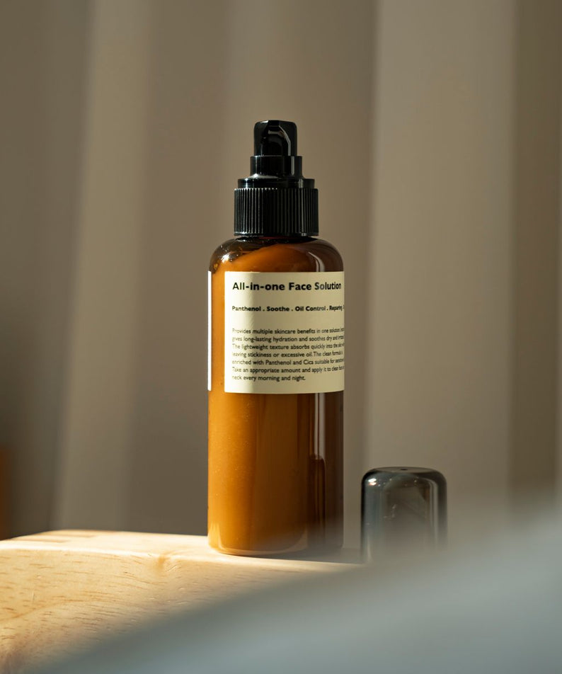 LAZY SOCIETY All-in-One Face Solution 150g, flacon pompe en verre ambré sur table bois, salle de bain, fond flou.