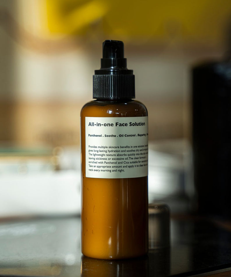 LAZY SOCIETY All-in-One Face Solution 150g, flacon brun à pompe sur table, arrière-plan flou, salle de bain.