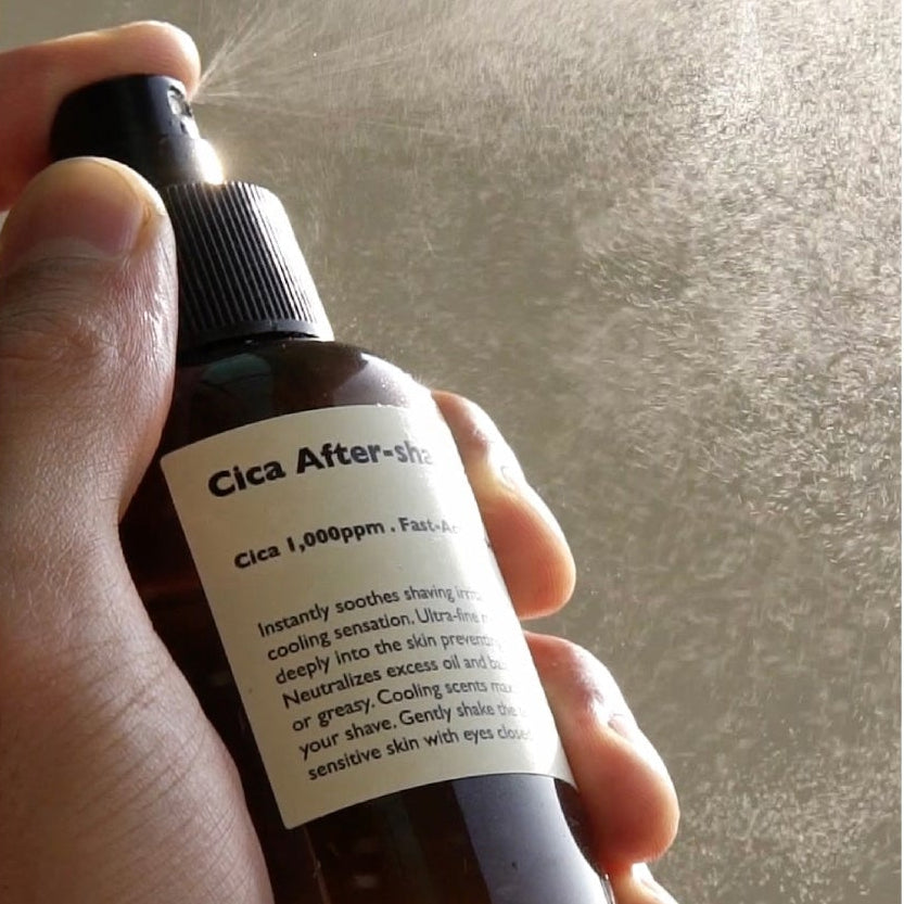 LAZY SOCIETY Cica Aftershave Toner Spray 120ml, main tenant le flacon sur fond texturé, étiquette lisible