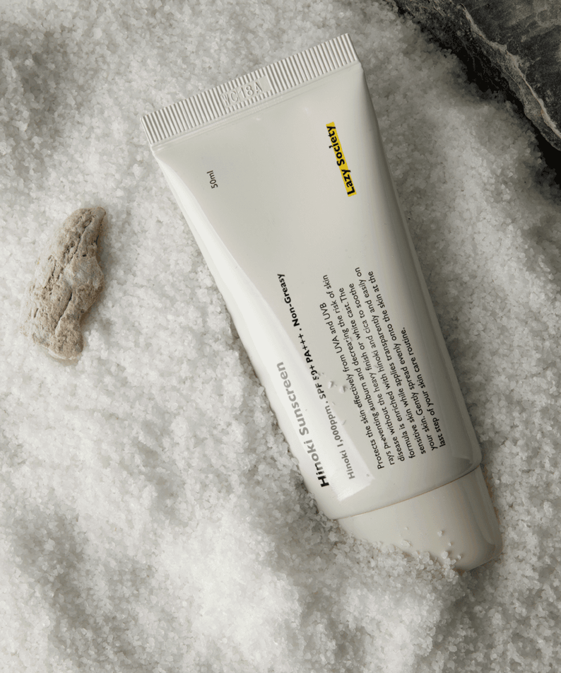 Décris l'image en français : LAZY SOCIETY Hinoki Sunscreen SPPF 50+ PA+++ 50ml, tube crème sur fond texturé, marque au visuel.