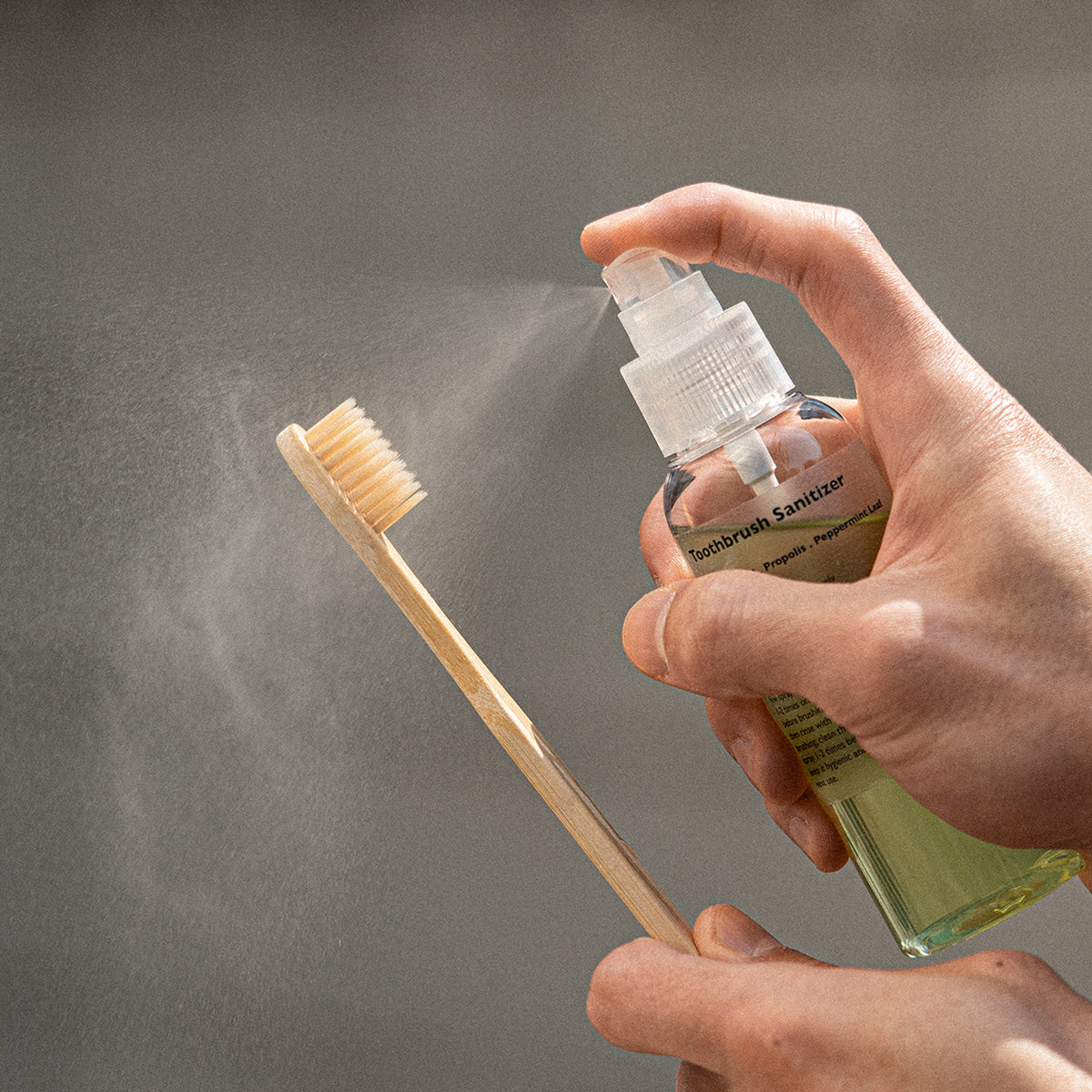 Décris l'image en français : LAZY SOCIETY Toothbrush Sanitizer 100ml, main vaporisant sur brosse, fond gris, marque visible.