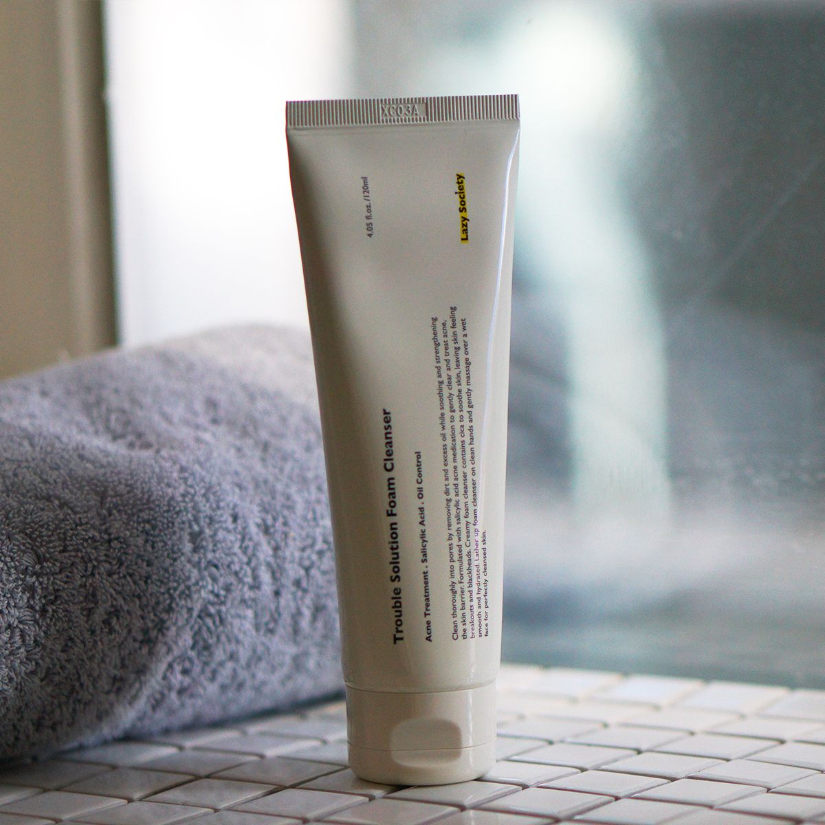 LAZY SOCIETY Trouble Solution Foam Cleanser 120ml, photo du tube sur salle de bain, serviette et décor minimaliste.