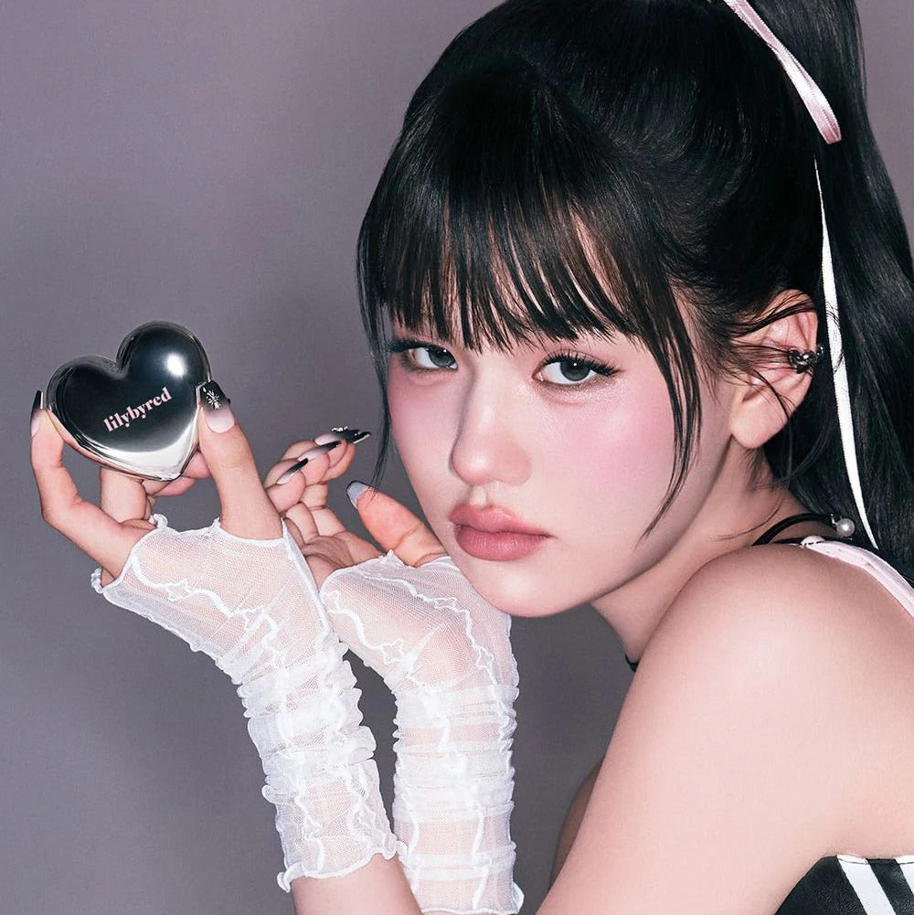 Décris l’image en français avec LILYBYRED Love Beam Cheek Mousse, marque sur un produit maquillage, pose beauté sur fond violet.