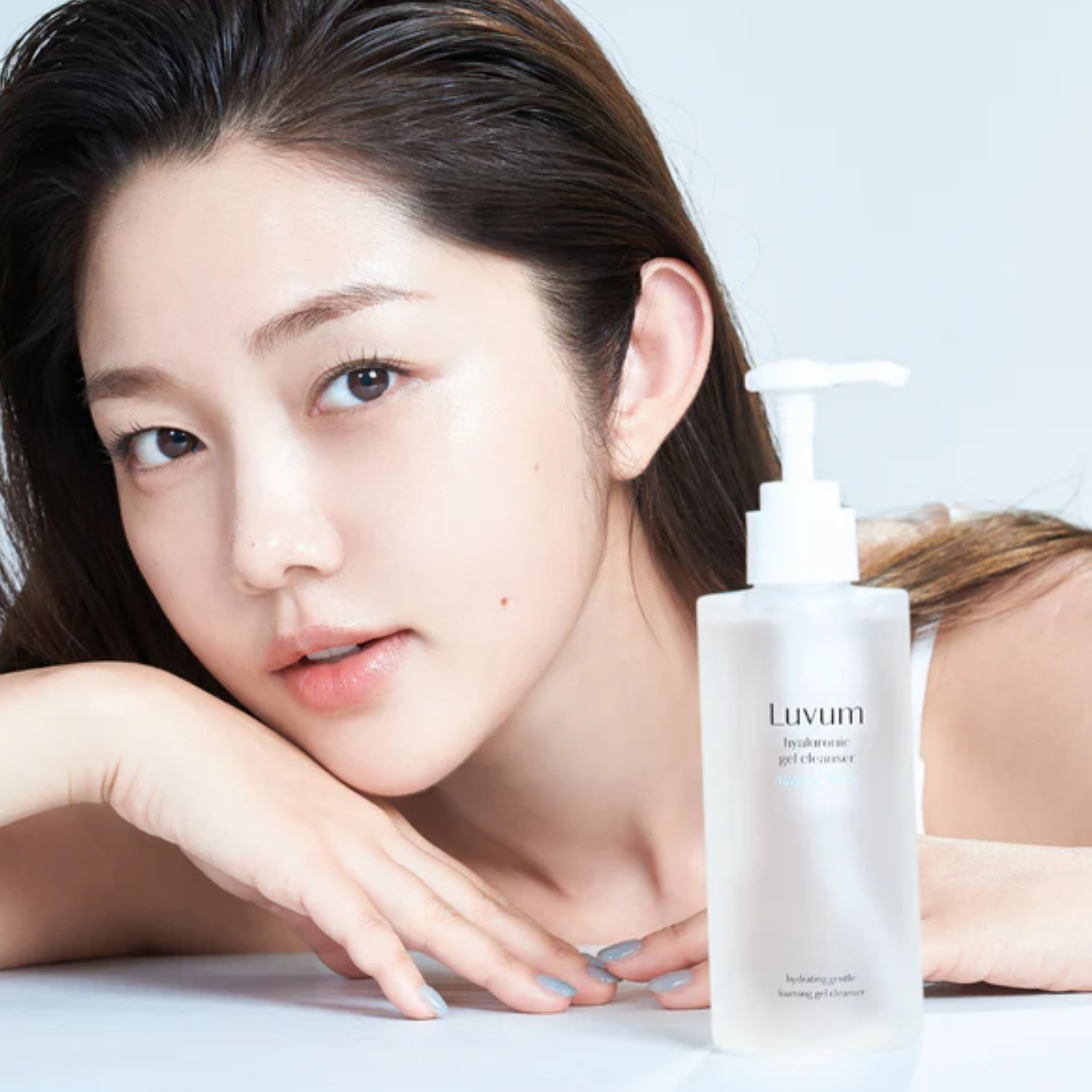 Décris l'image en français : LUVUM Bamboo Hyaluronic Acid Moisture Gel Cleanser 200ml, flacon blanc sur table, femme visage.