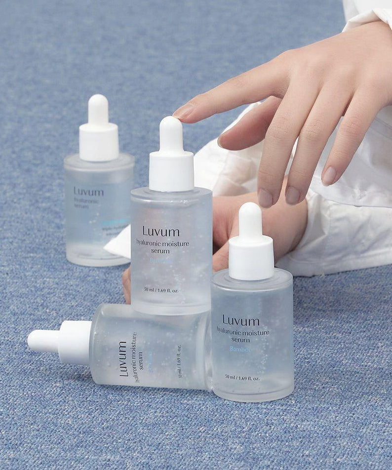 Décris l'image en français : LUVUM Bamboo Hyaluronic Acid Moisture Serum Set 50ml+50ml, flacons sur tissu bleu, main tenant le sérum