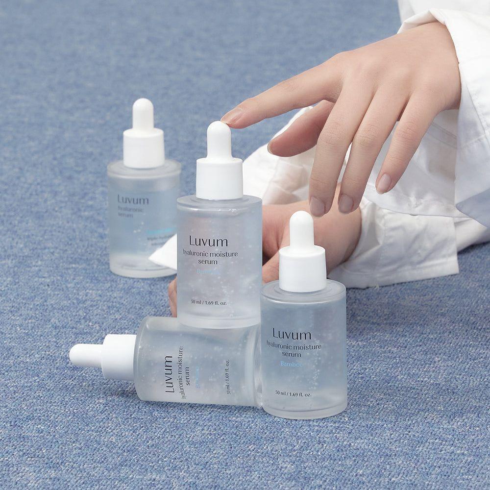 Décris l'image en français : LUVUM Bamboo Hyaluronic Acid Moisture Serum Set 50ml+50ml, flacons sur tissu bleu, main tenant le sérum