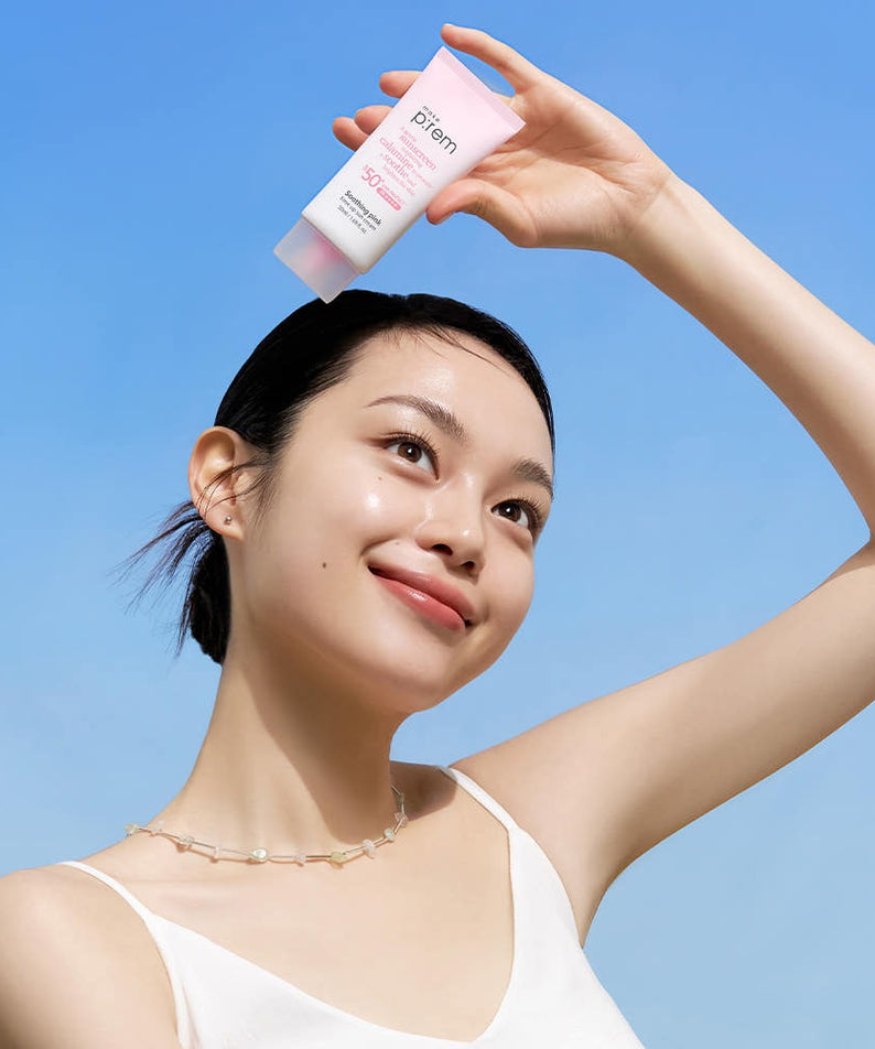 Femme souriante tenant MAKE P:REM Soothing Pink Tone-Up Sunscreen SPF 50+ PA++++ 50ml sous un ciel bleu, protection solaire efficace.