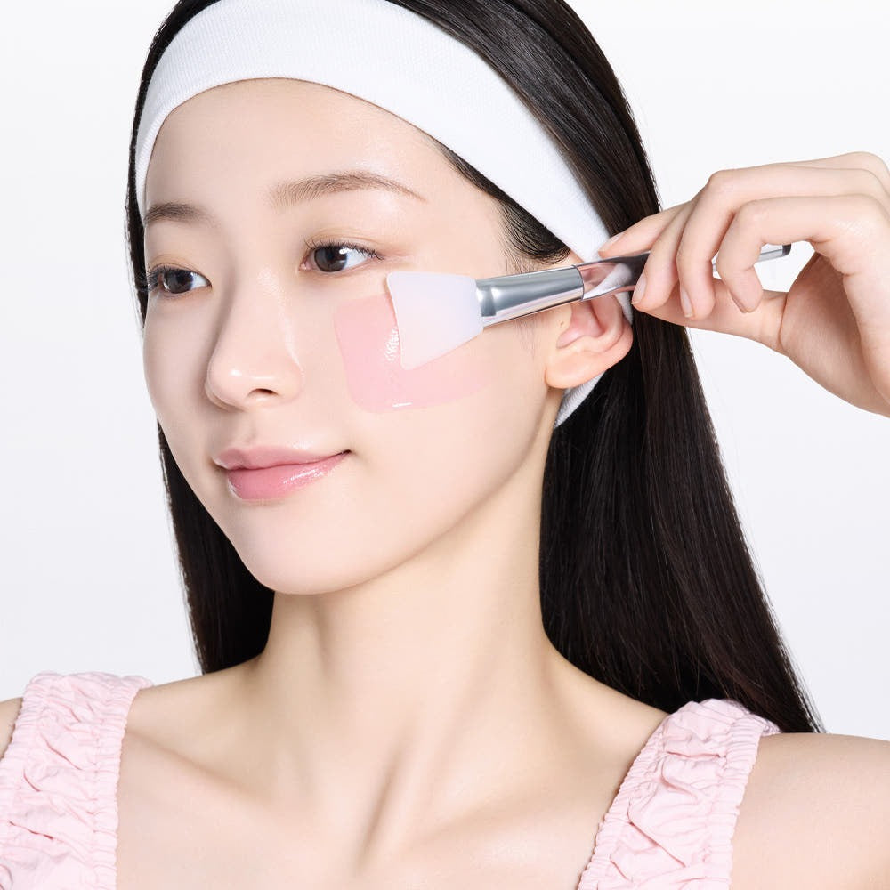 MAMONDE Flora Glow Rose Sleeping Mask 80ml, femme applique une crème rosée au pinceau sur le visage, fond blanc.