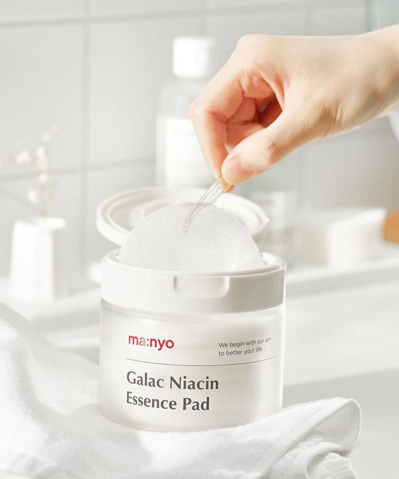 MANYO Galac Niacin Essence Pad 80 Sheets pour soin du visage, boîte ouverte avec main prenant un disque de coton.