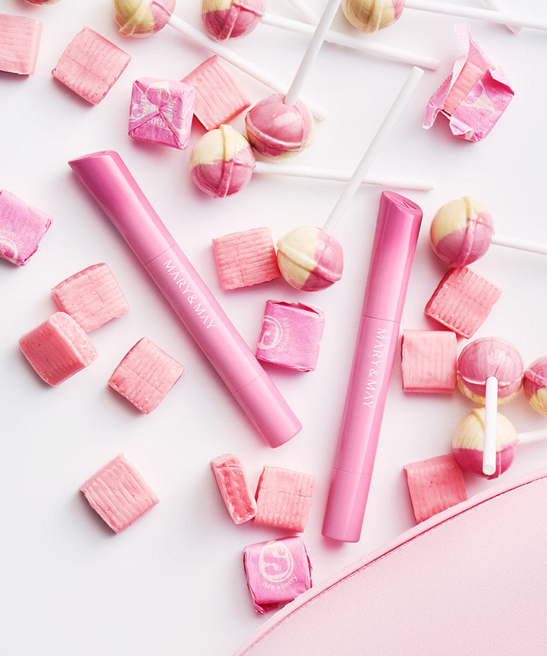 Décris l’image en français : MARY&MAY Blooming Glow Lip 09 Sunset Tangerine 1.7g, lèvres roses, bonbons sur fond clair.