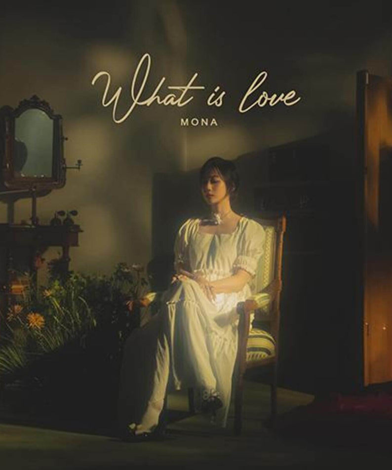 MONA - [What Is love] - Album Kpop : couverture sombre, artiste en robe blanche assise, studio rétro, fleurs et lumière chaude.