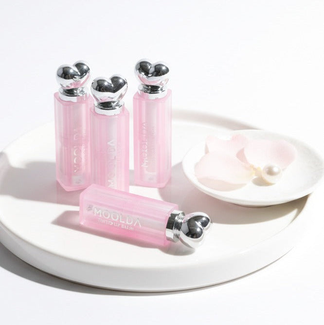 MOOLDA Tinted Lip Balm : tubes roses avec embouts argentés sur plateau blanc, contexte maquillage, marque MOOLDA.