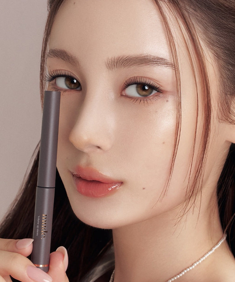 Décris l'image en français : MUDE Mood Volume Up Skinny Mascara, main tenant le mascara sur un visage maquillé.