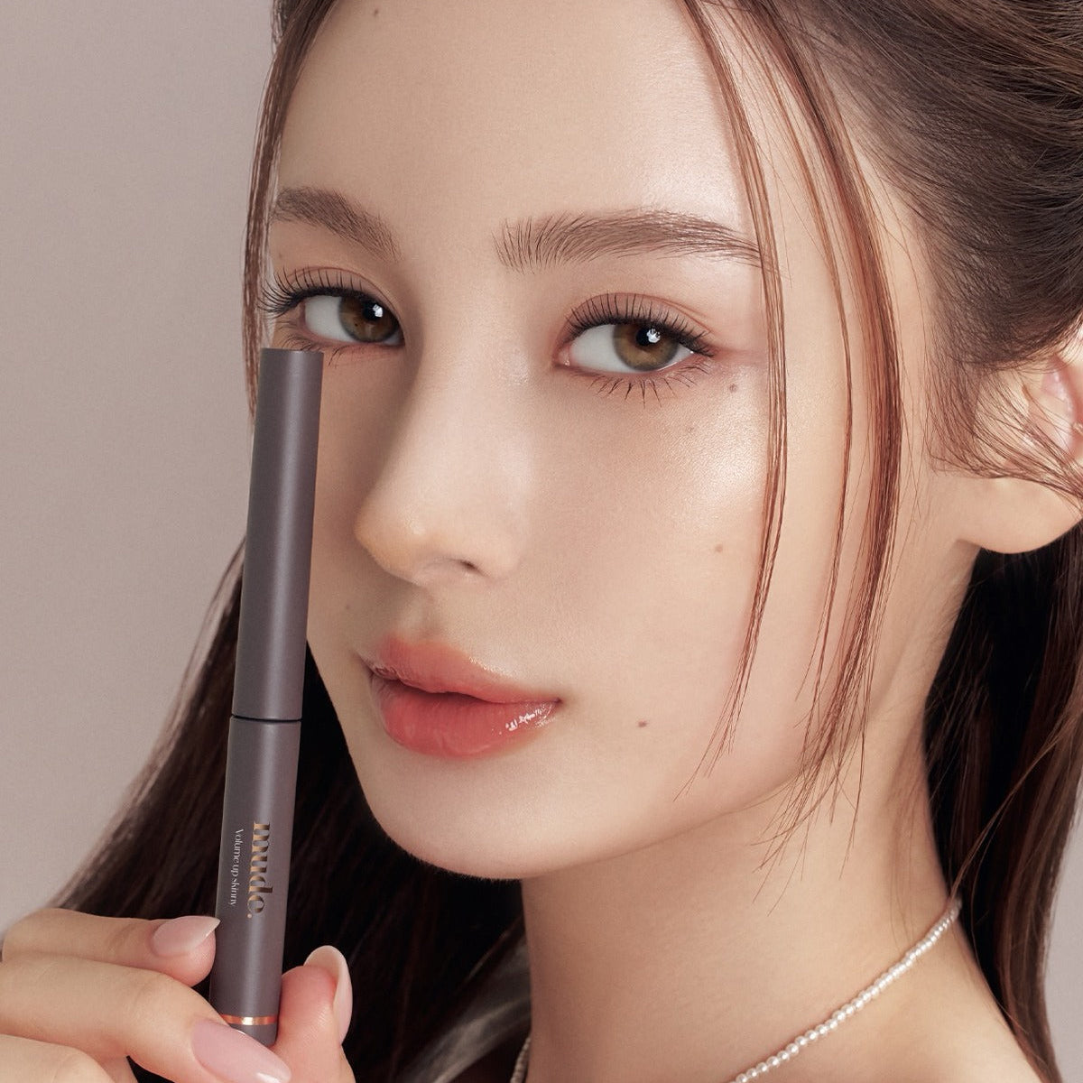 Décris l'image en français : MUDE Mood Volume Up Skinny Mascara, main tenant le mascara sur un visage maquillé.
