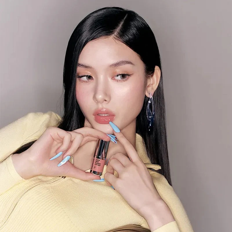 NAMING Overdew Glossy Lip Tint, femme aux lèvres brillantes tenant le produit sur fond neutre, maquillage glam