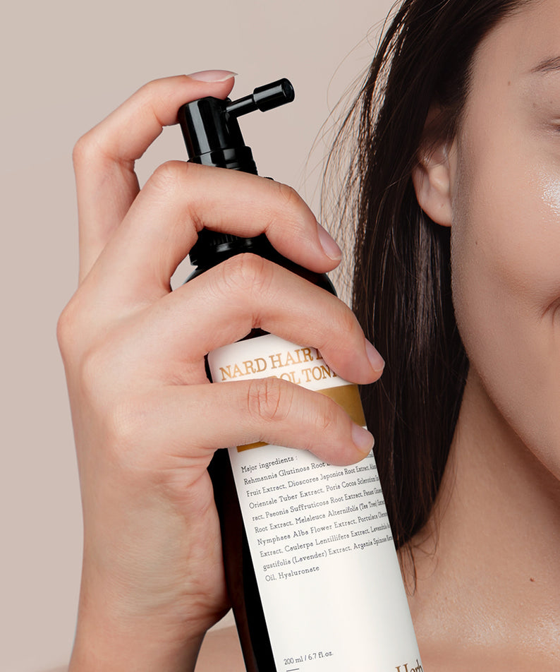 Décris l'image en français : NARD Hair Loss Relief Hair Tonic 200ml de la marque NARD, appliqué sur le visage, main tenant le flacon.