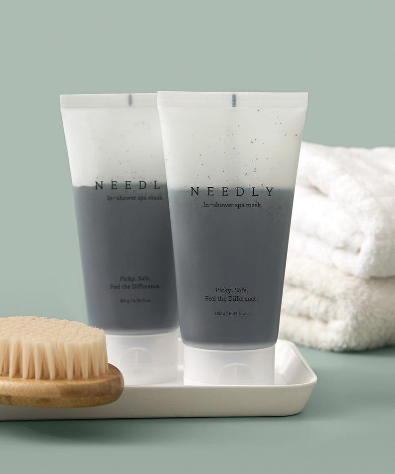 Deux tubes de NEEDLY Ishower Spa Mask 180g, brosse à sec et serviettes blanches en arrière-plan, soin de la peau luxueux en douche.