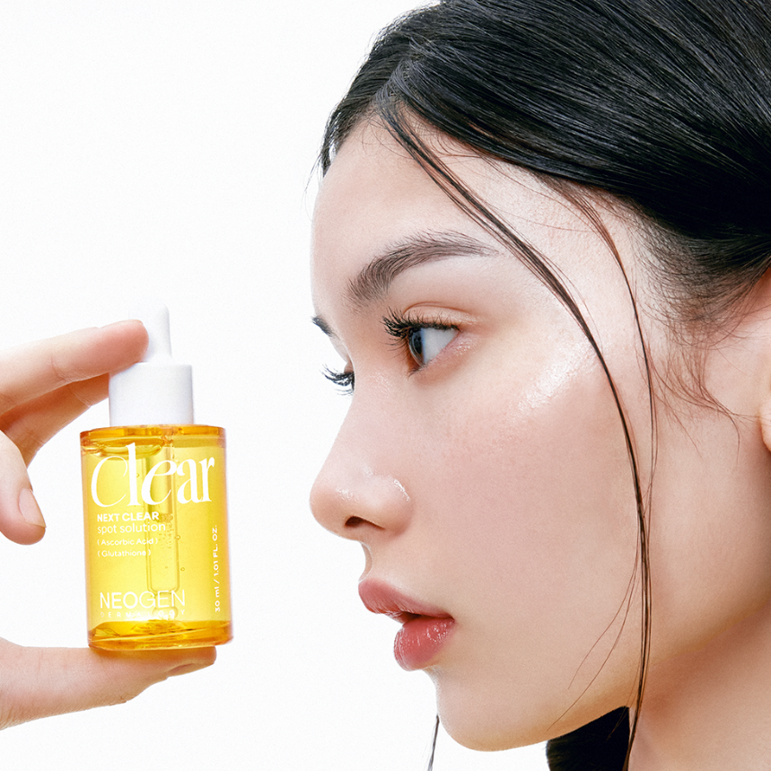 NEOGEN  Next Clear Spot Solution 32ml, flacon jaune de la marque NEOGEN près d’un visage féminin sur fond blanc.