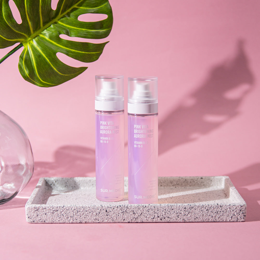 NEOGEN Pink Vita Aurora Mist 115ml de la marque NEOGEN, flacons roses sur support gris, fond rose avec feuilles tropicales.
