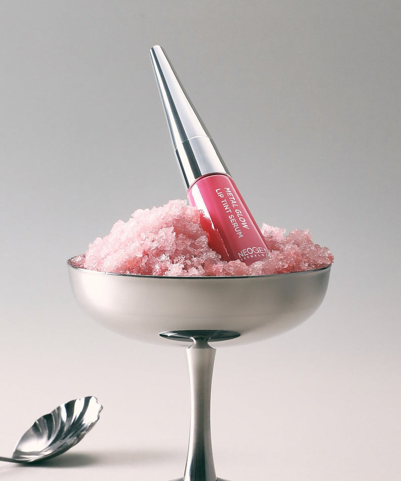 NEOGEN Metal Glow Lip Tint Serum, flacon sur un support argent avec texture rosée, fond clair minimaliste.