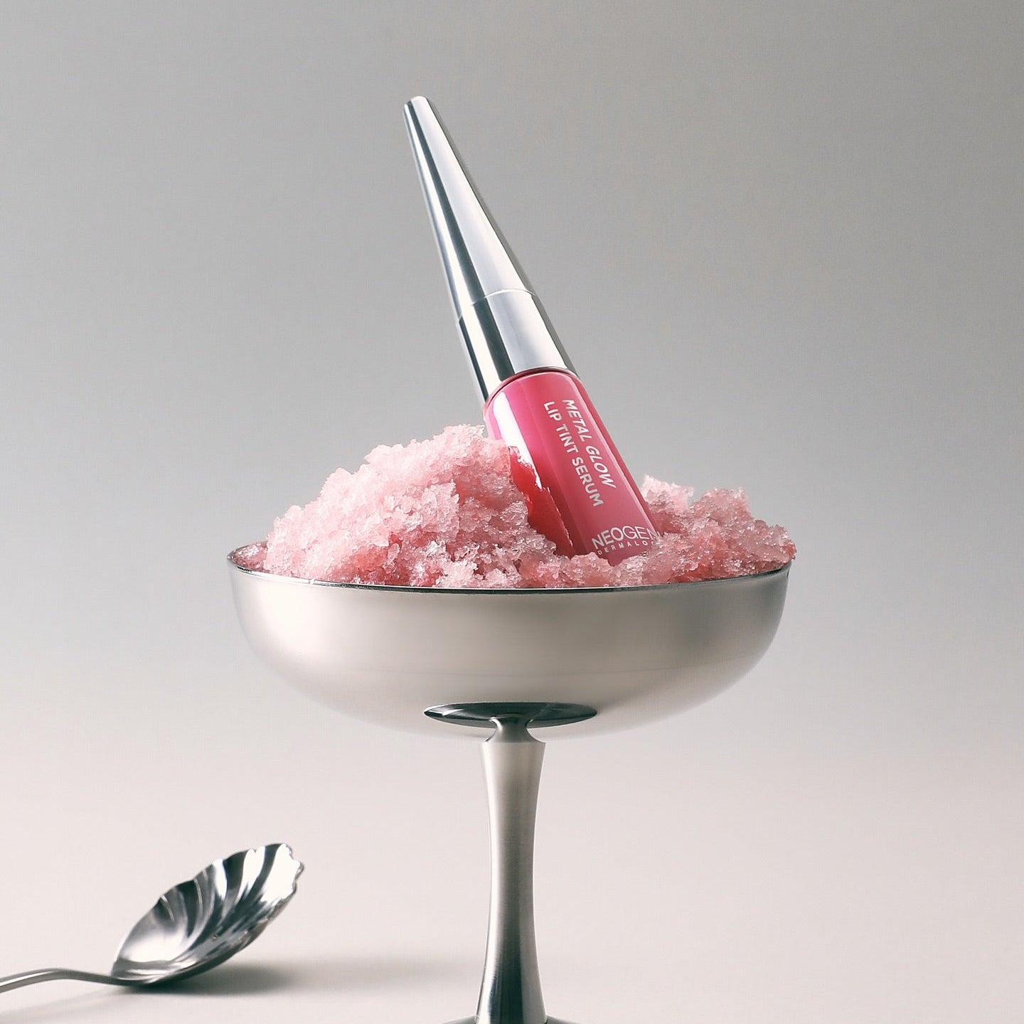 NEOGEN Metal Glow Lip Tint Serum, flacon sur un support argent avec texture rosée, fond clair minimaliste.