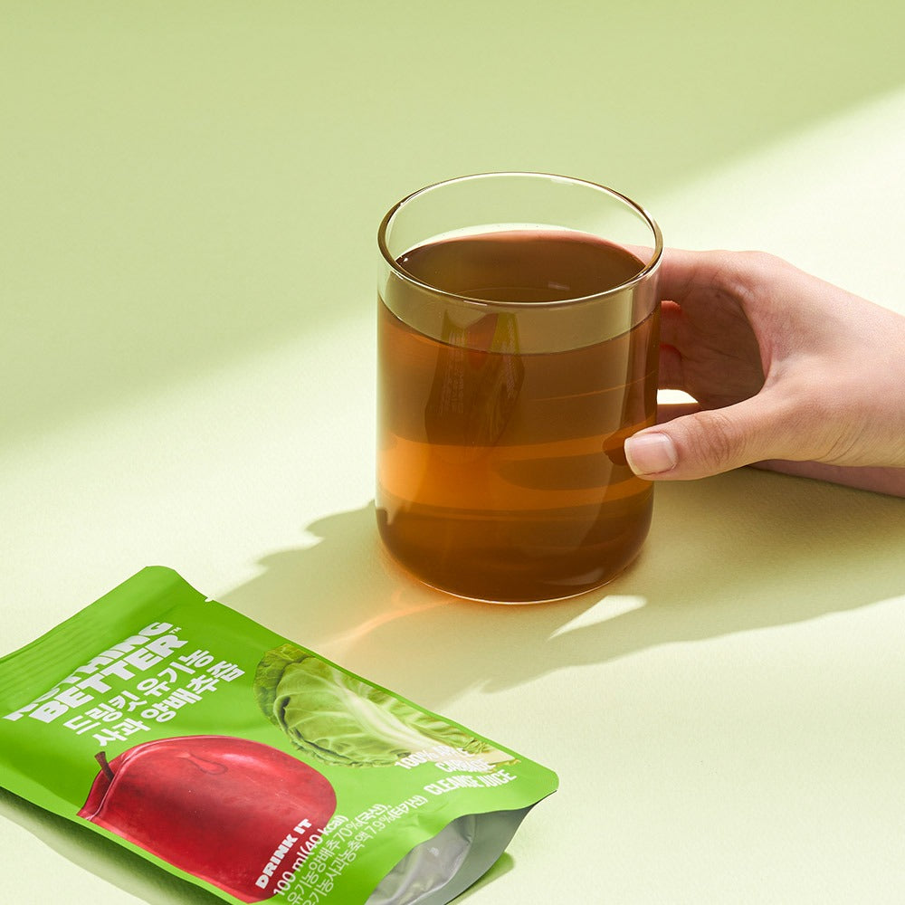 Décris l'image en français : NOTHING BETTER Drinkit Organic Applr Cabbage Juice 100ml 24 Packets, mains tient mug, sachet vert.