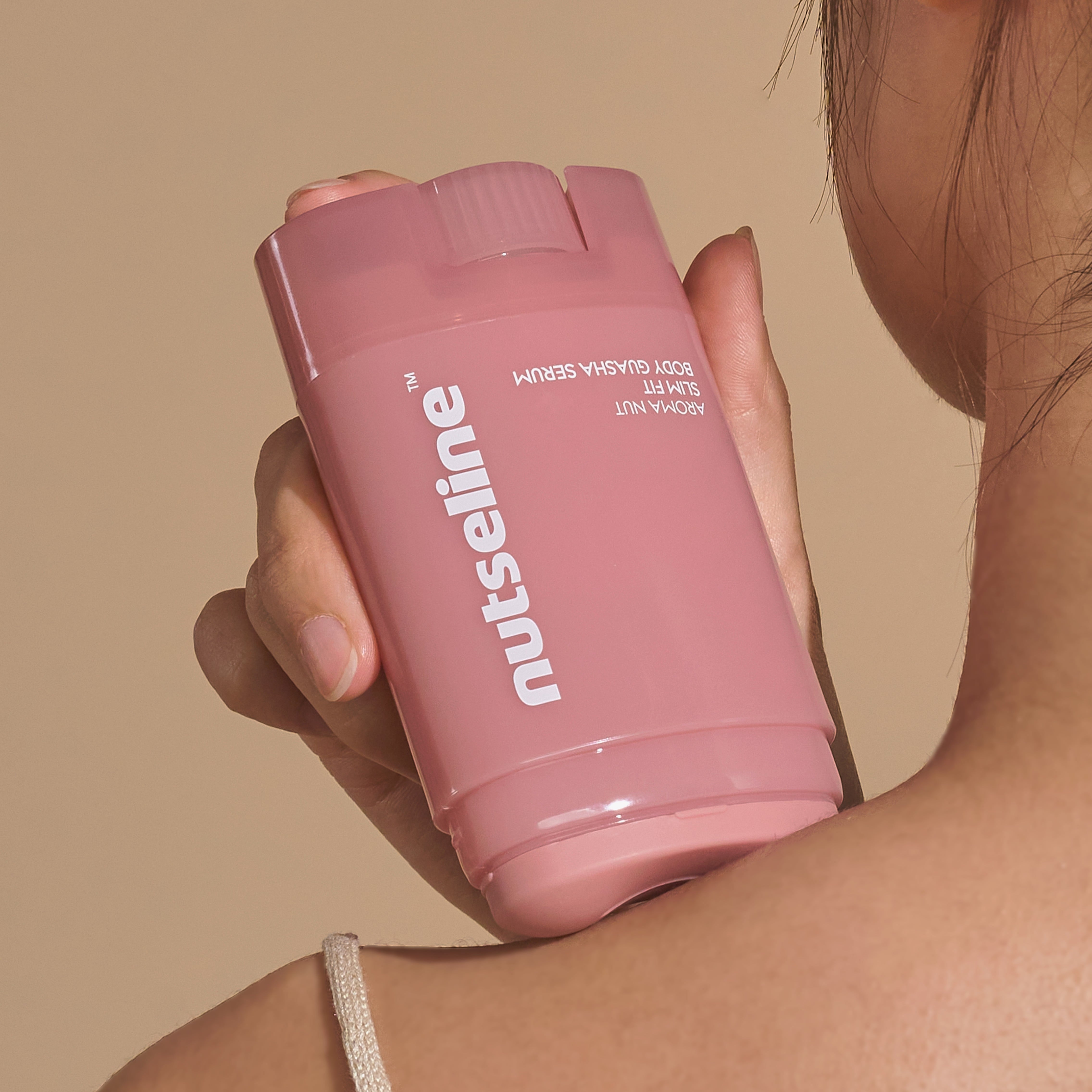 NUTSELINE Aroma Nut Slim Fit Body Guasha Serum 60ml, flacon rose tenu sur peau, contexte beauté en studio.