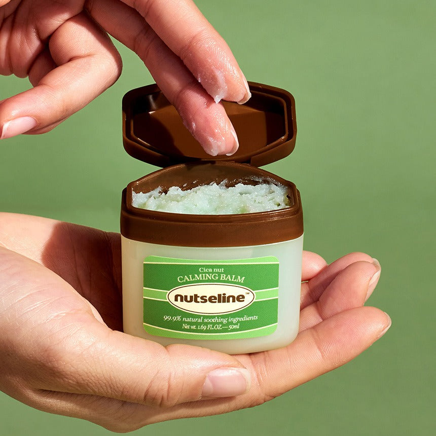 NUTSELINE Cica Nut Calming Balm Season 2 50ml : pot ouvert avec baume, mains, fond vert, contexte soins visage.