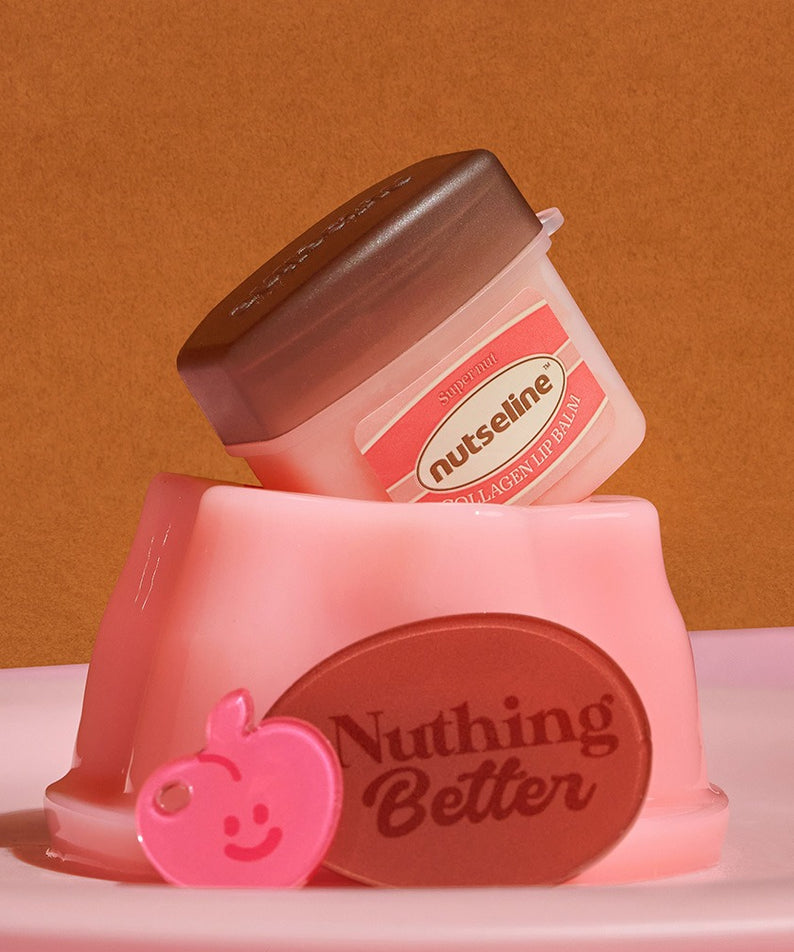 Le NUTSELINE Super Nut Collagen Keyring Lip Balm 11ml posé sur un support rose avec un charm pomme souriante et texte "Nuthing Better".