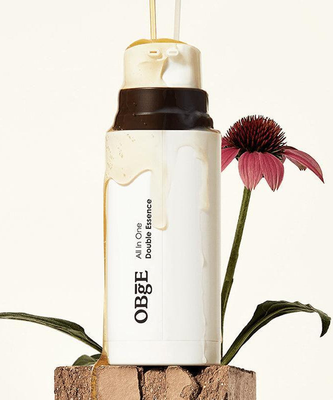 OBGE All-in-one double essence 100ml, bouteille cosmétique OBGE sur pierre avec fleur rouge, fond clair (photo produit)