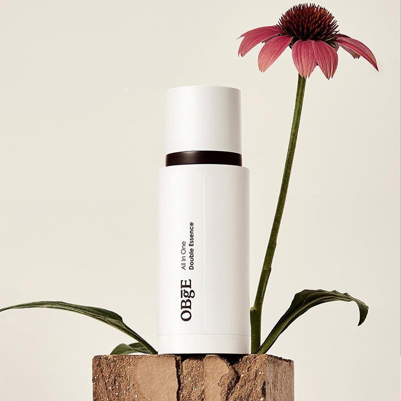 OBGE All-in-one double essence 100ml : flacon blanc OBGE sur socle bois, fleur violette en décor naturel, fond clair.