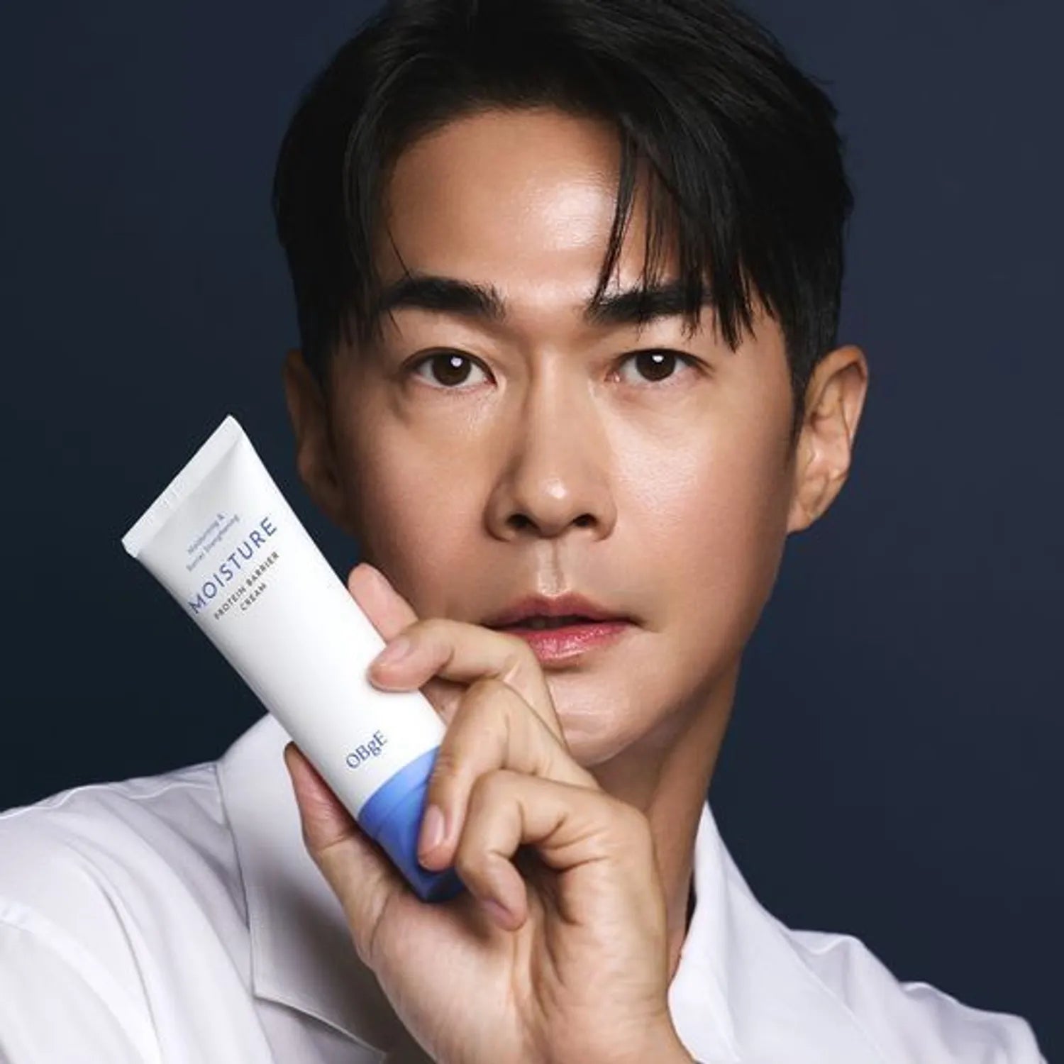 OBGE Moisture Protein Barrier Cream 150ml, tube blanc, présenté par un homme en studio sur fond bleu foncé.
