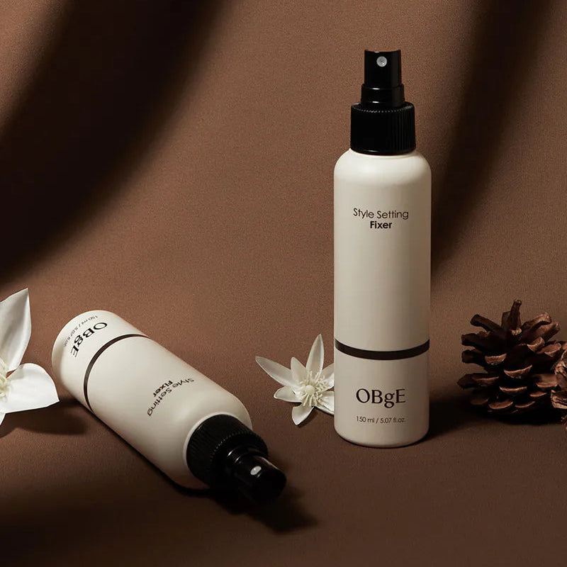 OBGE Style Setting Fixer 150ml, brume coiffante OBGE sur fond beige, avec fleurs et accessoires décoratifs.